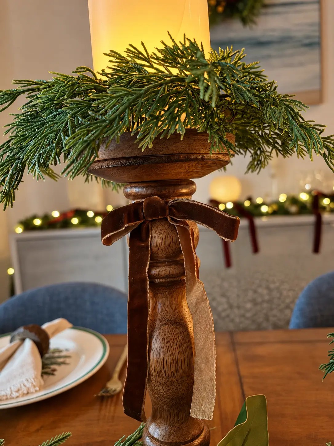 One turned wood candle holder dressed with a velvet bow and a cedar collar, its pillar candle lit, the place settings soft behind it