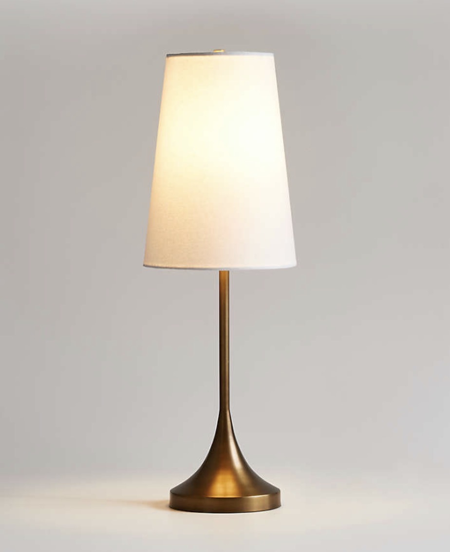 Brushed brass table lamp with a slim stem, flared base and tall white linen shade, lit