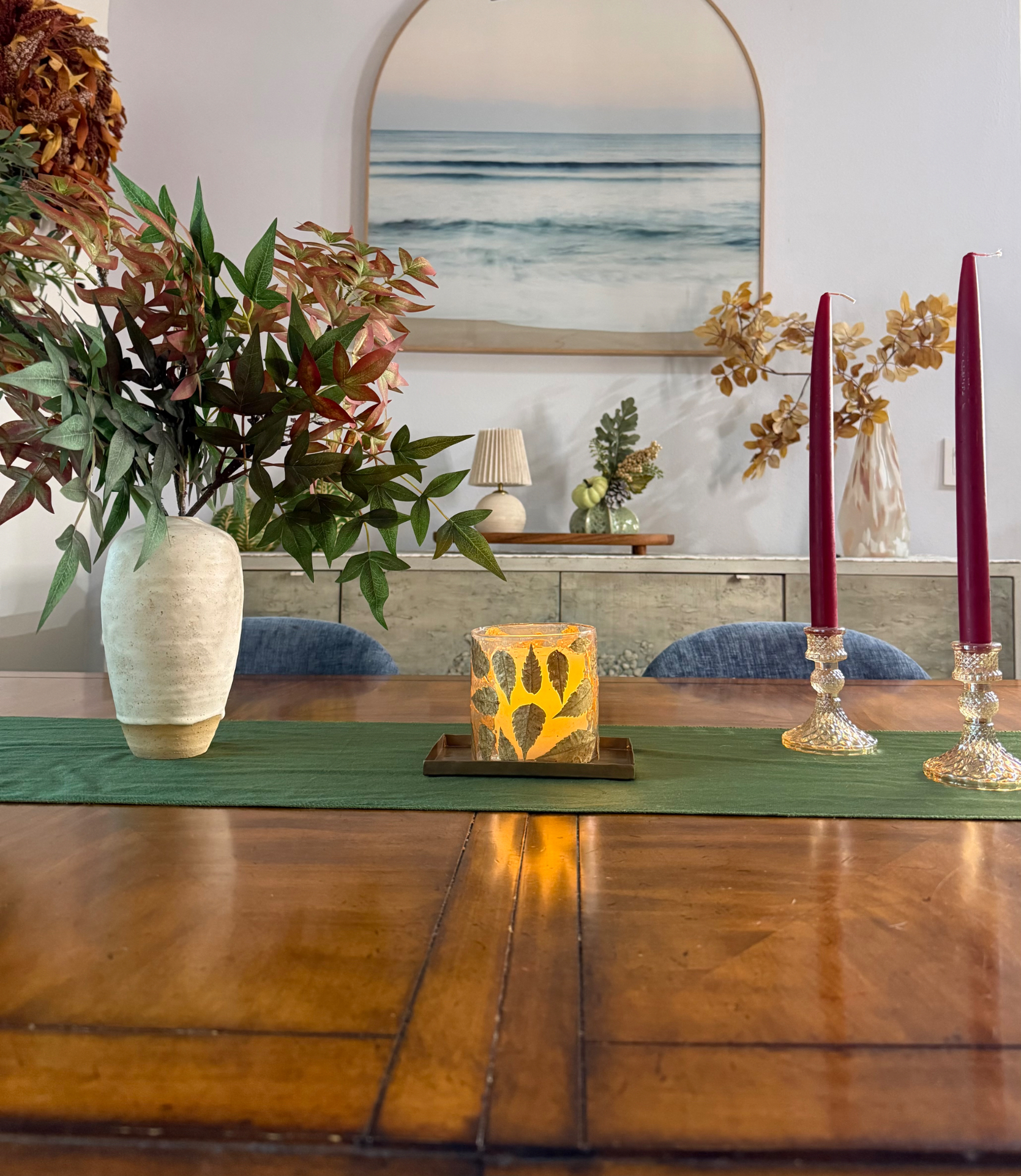 Golden Hour edit styled on a dining table: cream vase with autumn branches, glowing candle on a brass tray, and burgundy tapers in amber glass candleholders on a green runner