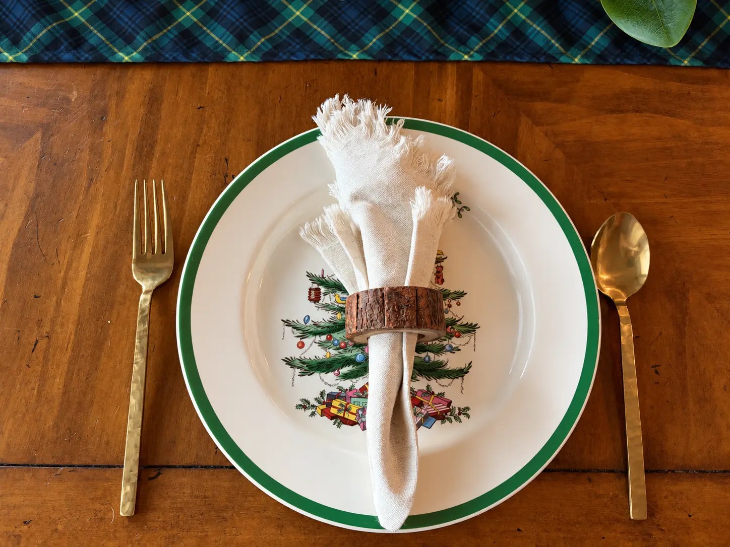 A single place setting from above: a white plate with a green border, a natural-fringe napkin slipped through a bark-edge ring, and hammered brass flatware at the edge of the tartan runner