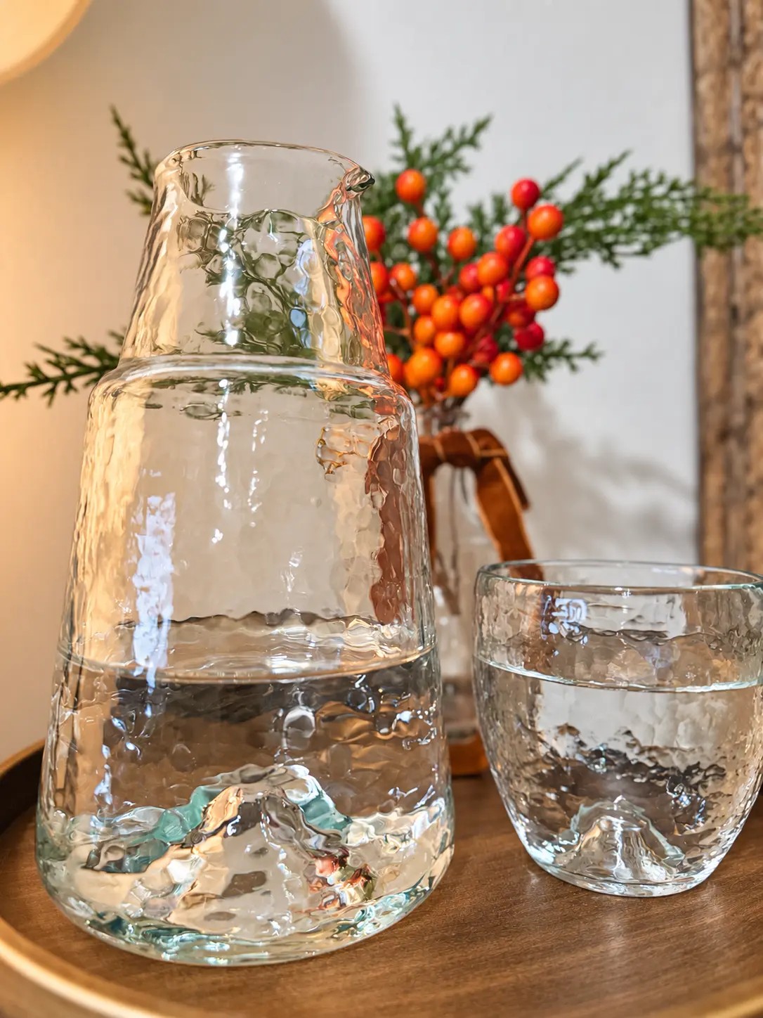 A pebbled recycled-glass carafe and its matching drinking glass, both filled with water, standing side by side on a round wooden tray