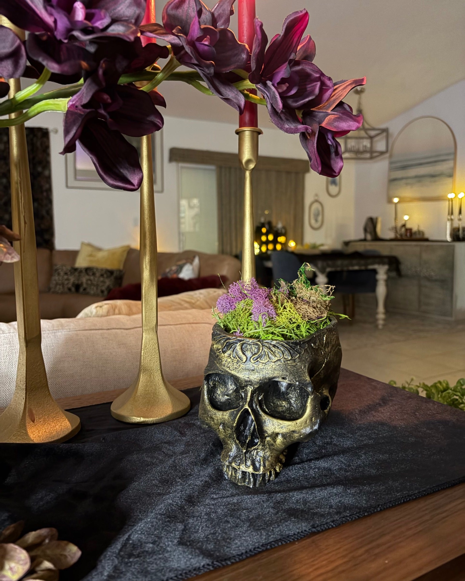 Close-up of the moss-filled antique-brass skull planter on black velvet beneath plum orchids and brass candlesticks