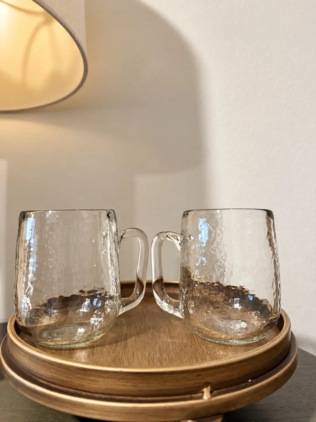 Two empty pebbled recycled-glass mugs side by side on a tiered brass-toned tray, lamplight warm behind them